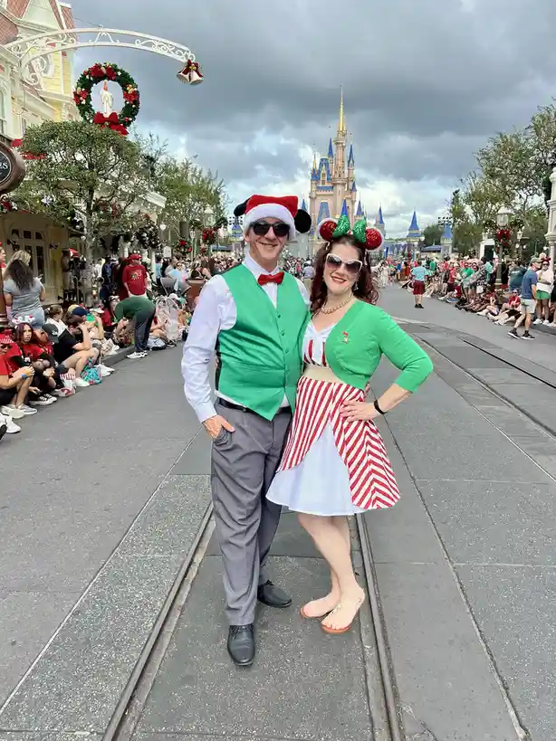 A man and woman pose for a photo on tram tracks at a Disney park, with the castle in the background. The man wears a green…