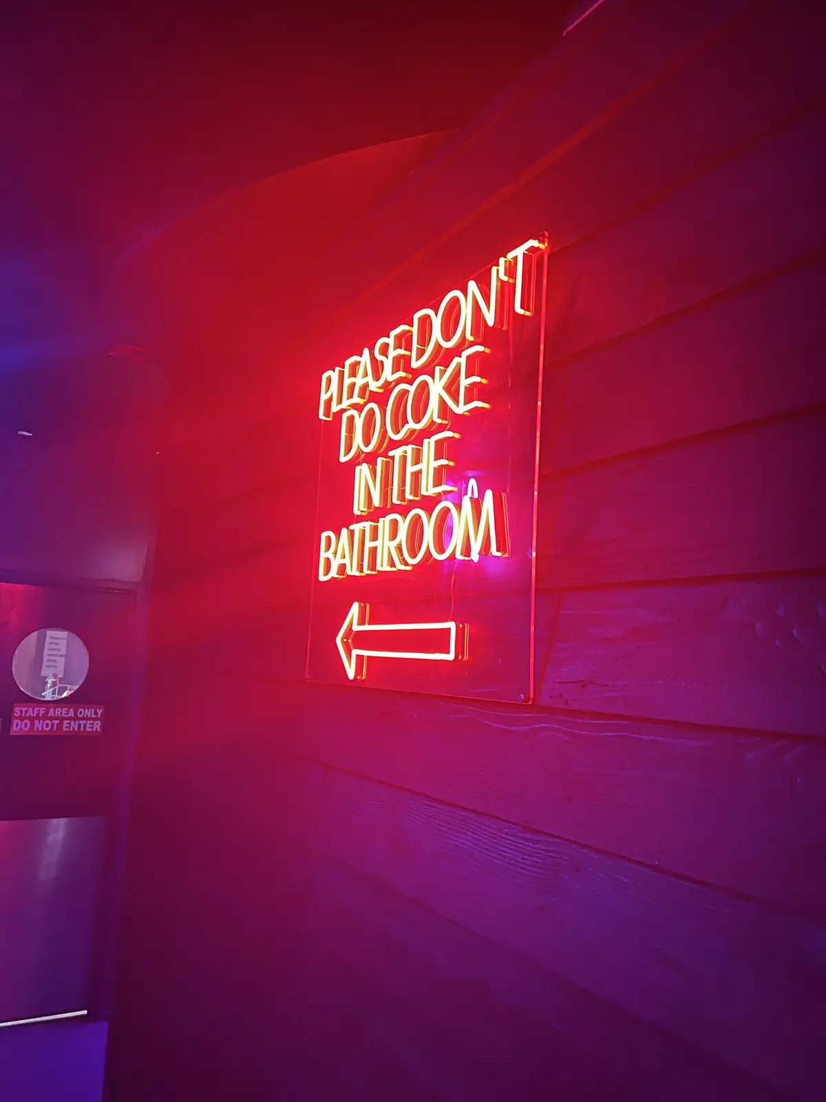 A red neon sign mounted on a wooden wall reads 'PLEASE DON'T DO COKE IN THE BATHROOM' with an arrow pointing left. To the…