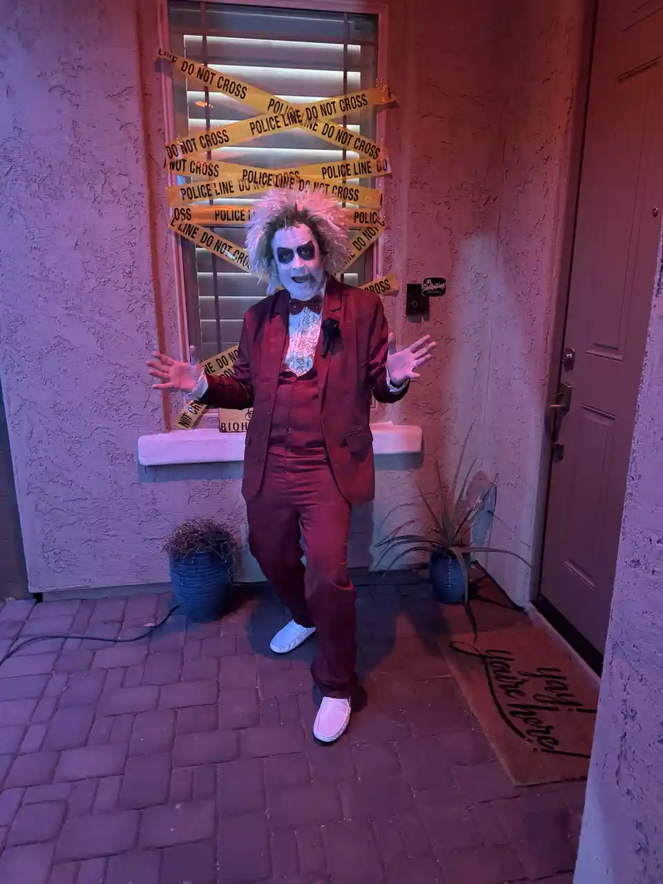 A person dressed as Beetlejuice stands in front of a house entrance with arms outstretched, surrounded by crisscrossing…