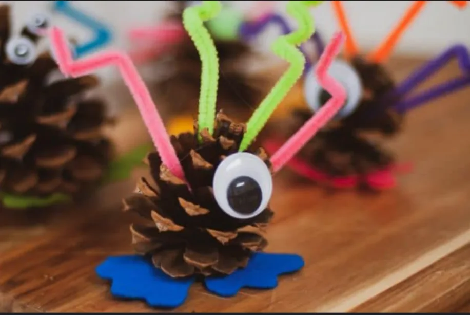 A close-up photo shows a pinecone decorated as a monster with a large googly eye, colorful pipe cleaner limbs, and a blue…