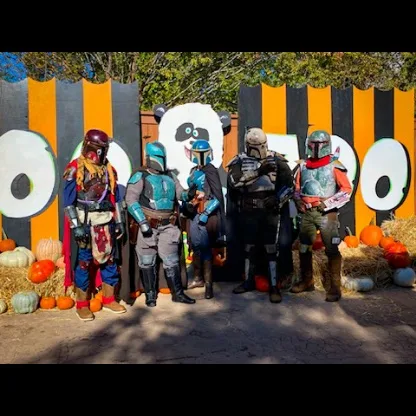 Five people dressed as Star Wars Mandalorian characters stand in front of a Halloween-themed backdrop with large cartoon…