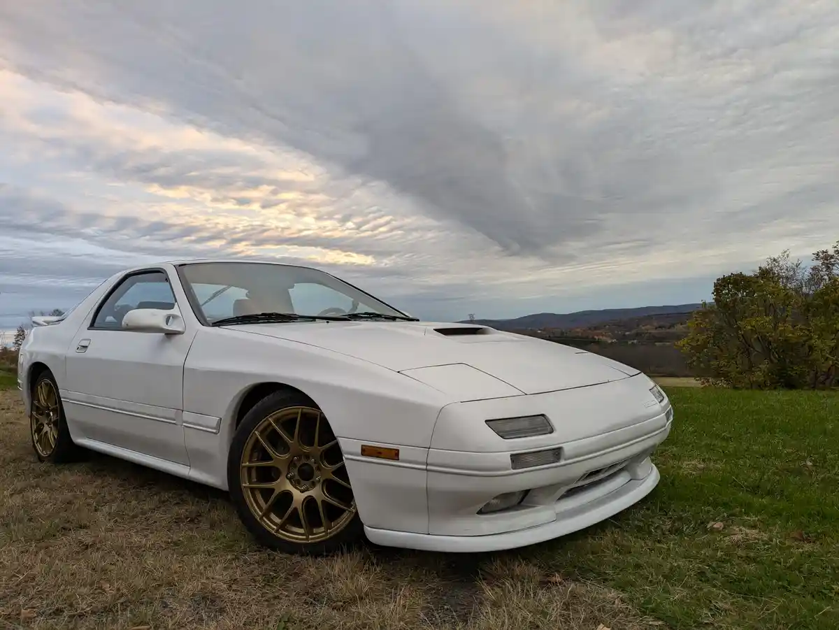 A white 1980s-era sports car, likely a Mazda RX-7, is parked on grass under a dramatic, cloudy sky with autumn trees in the…