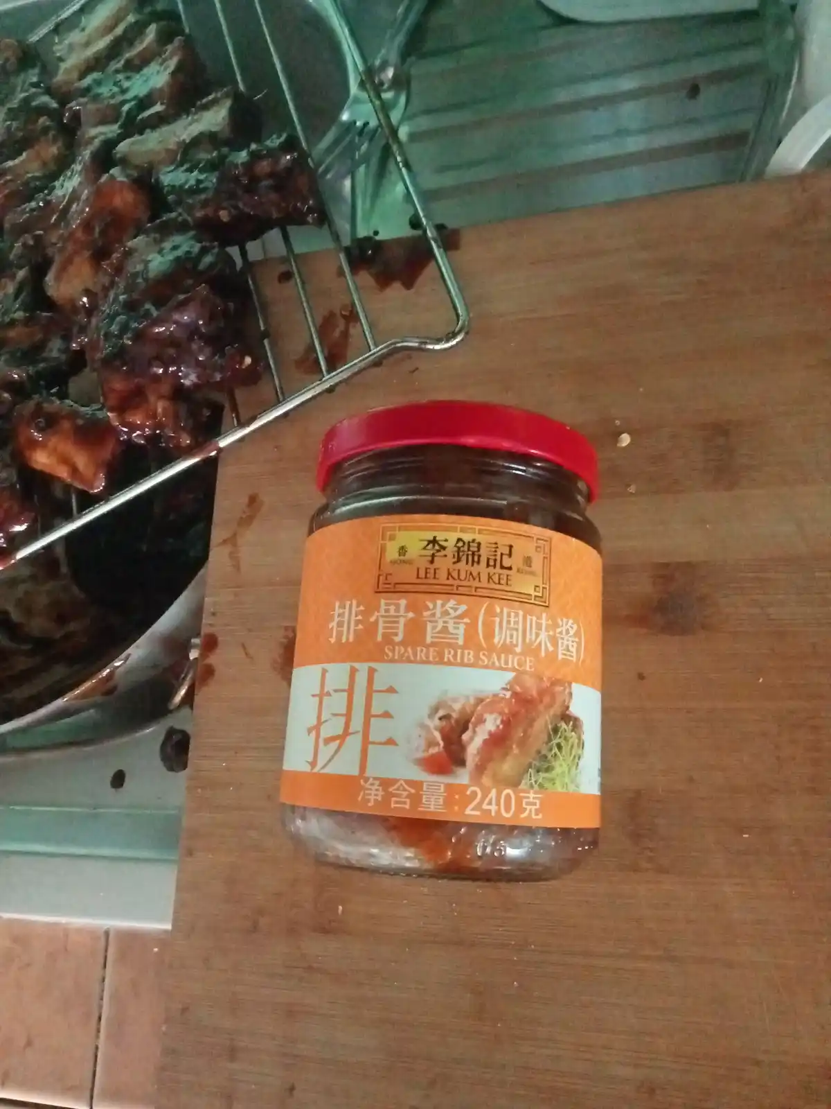 A jar of Lee Kum Kee Spare Rib Sauce sits on a wooden surface next to a metal rack holding cooked ribs that have a dark…
