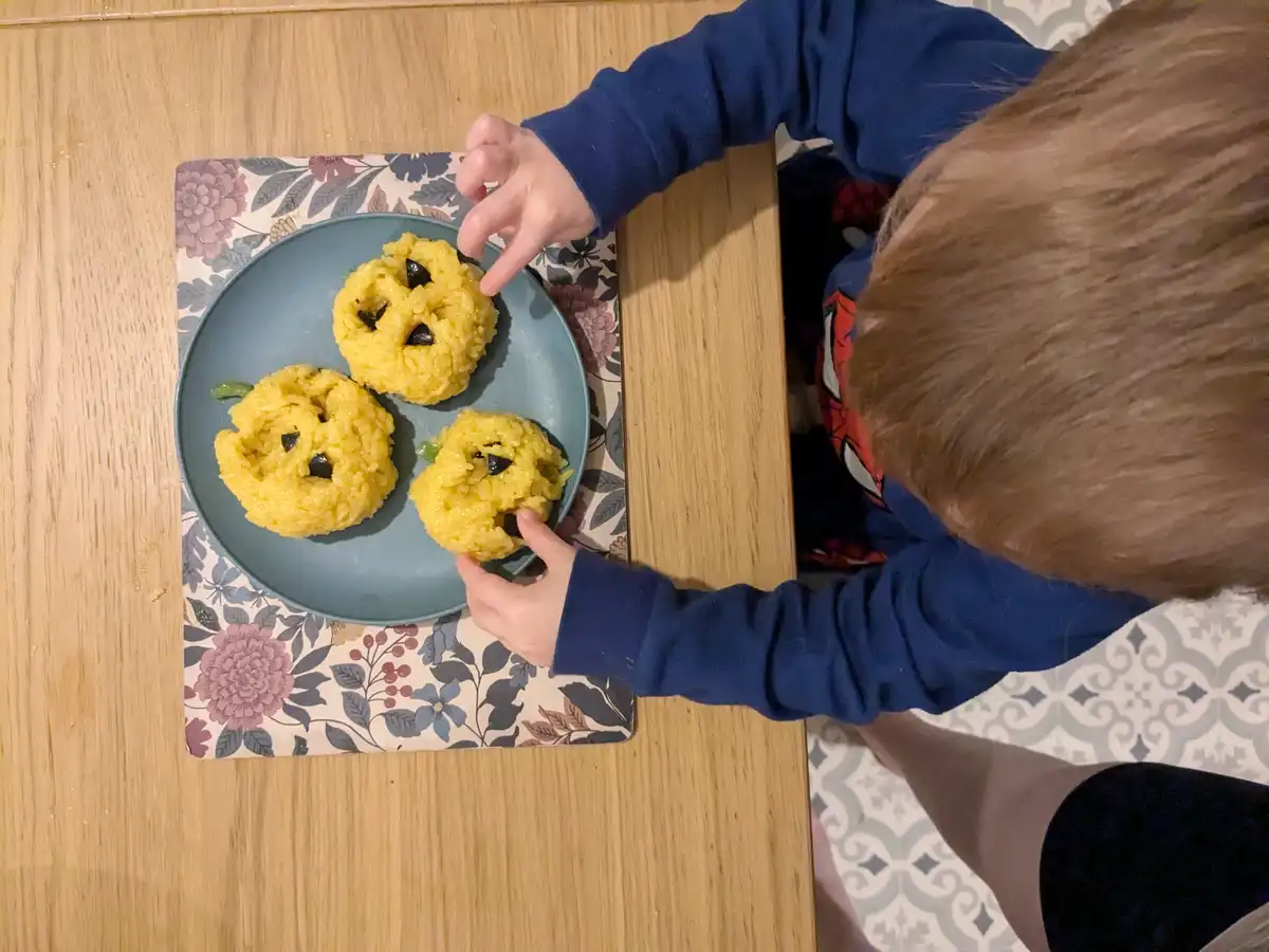A child in a blue sweatshirt is reaching for three yellow rice balls shaped like pumpkins with black olive eyes and green…