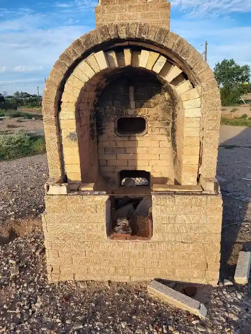 An outdoor brick oven with a large arched opening, showing signs of disrepair and abandonment, sits on gravel under a partly…