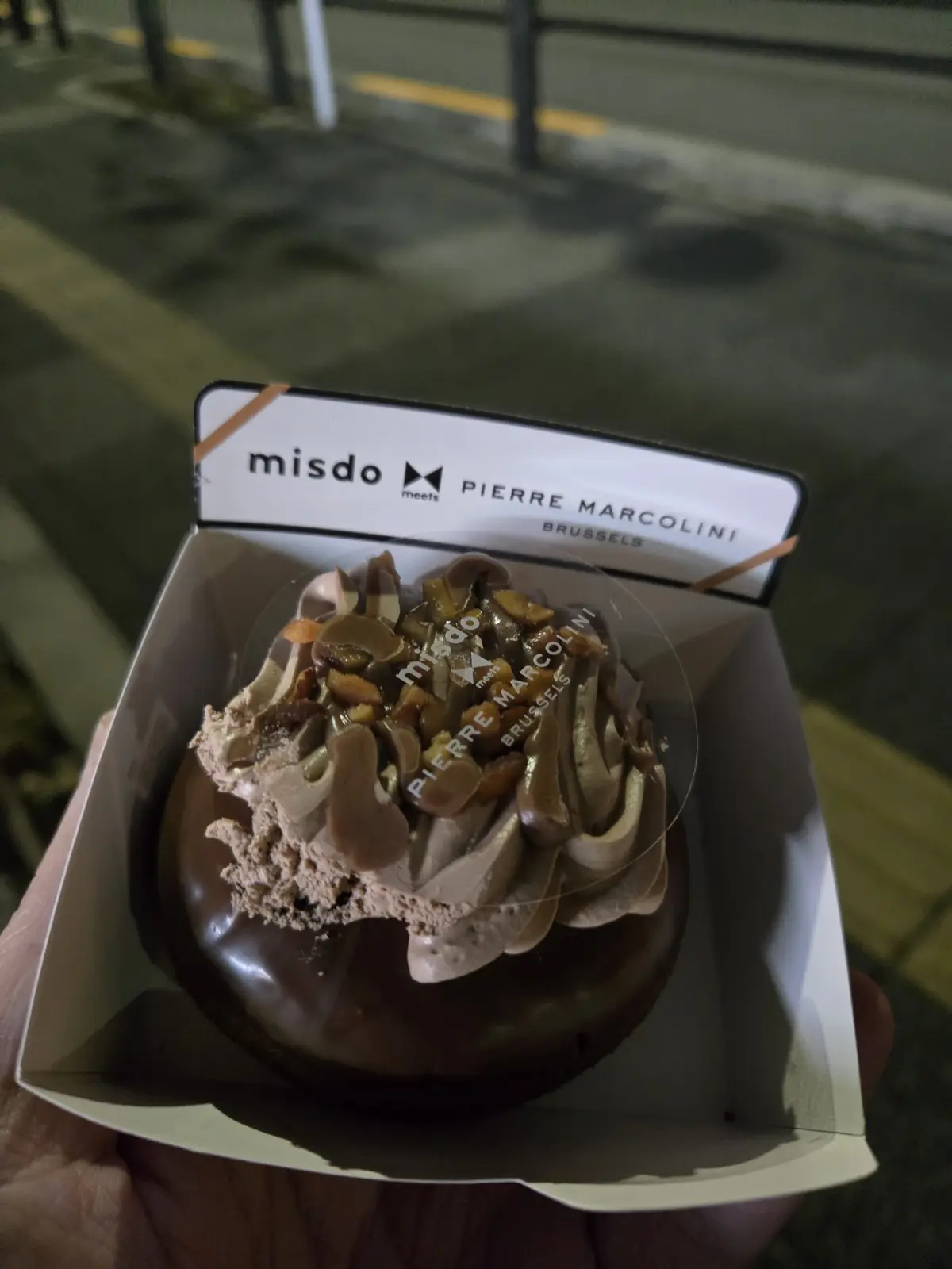 A hand holds a chocolate dessert in a branded box labeled 'misdo meets PIERRE MARCOLINI BRUSSELS'. The dessert has chocolate…