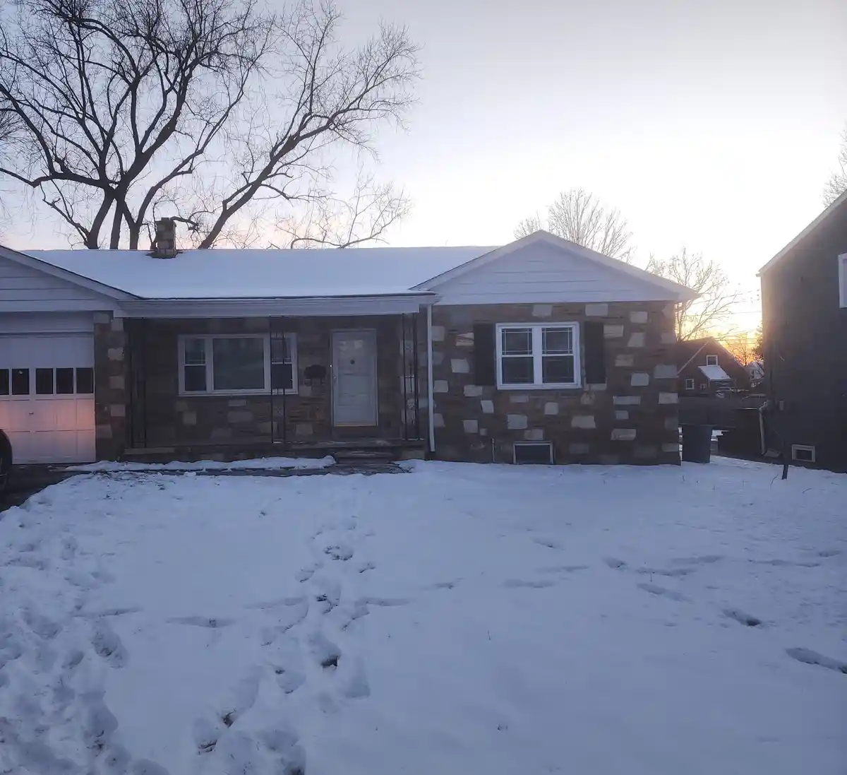 A snowy front yard leads to a stone-faced single-story house with a white door and windows, under a pale sky with bare…
