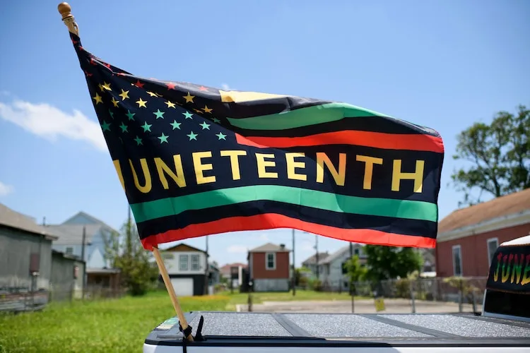 A Juneteenth flag, featuring stars and stripes with green, black, and red horizontal bars and the word 'JUNETEENTH' in bold…