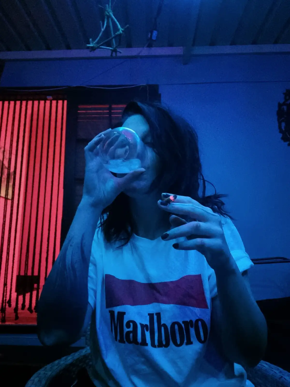 A person with dark hair is shown drinking from a glass while holding a lit cigarette in their other hand. They are wearing a…