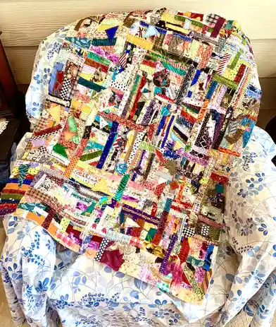 A colorful, patchwork quilt made from numerous small, varied fabric scraps is draped over a chair covered in a blue floral…