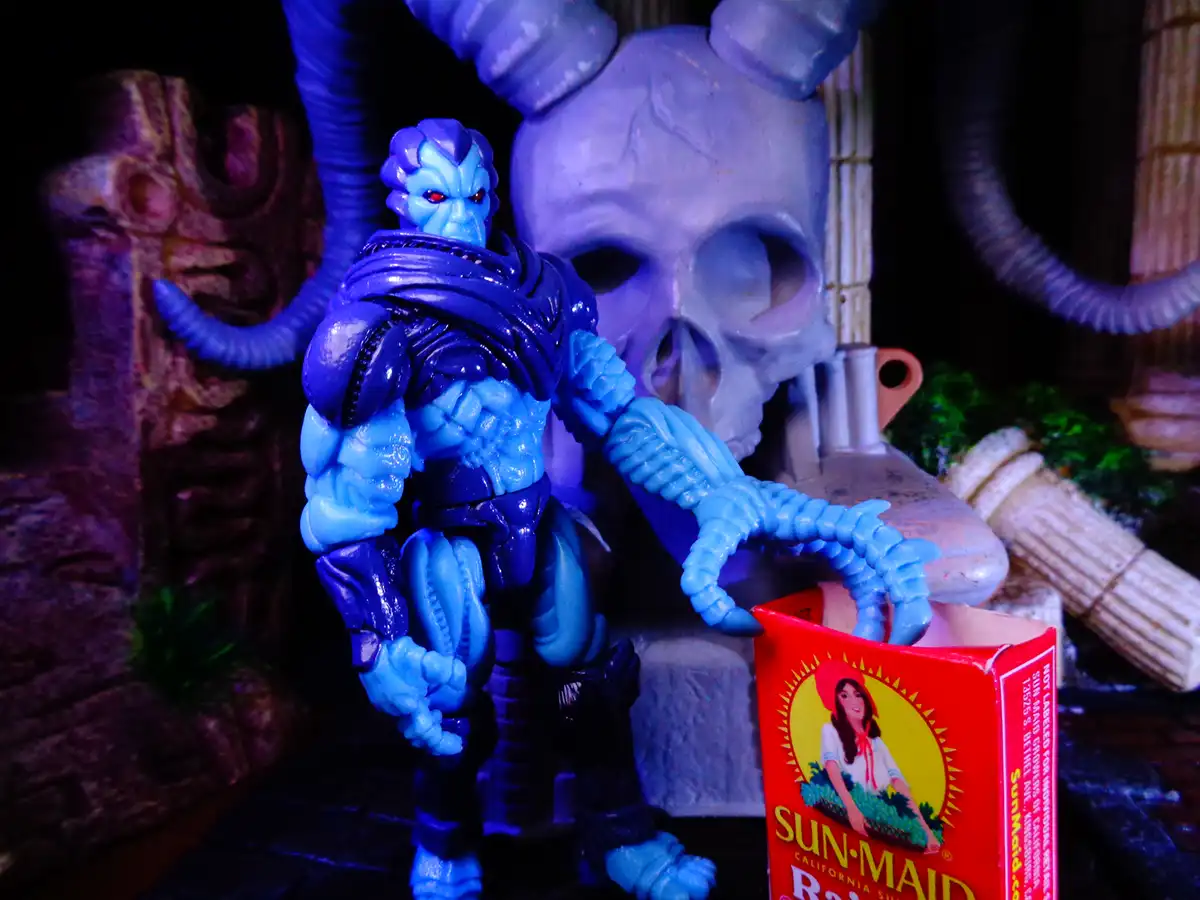 A blue, muscular, horned demon action figure with red eyes stands in a dark, skull-themed diorama, holding a red Sun-Maid…