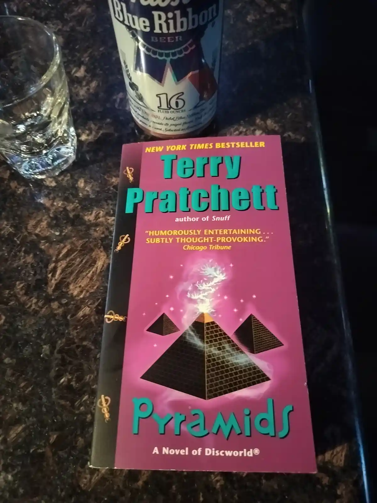 A book titled 'Pyramids' by Terry Pratchett, part of the Discworld series, sits on a dark countertop next to a can of Blue…