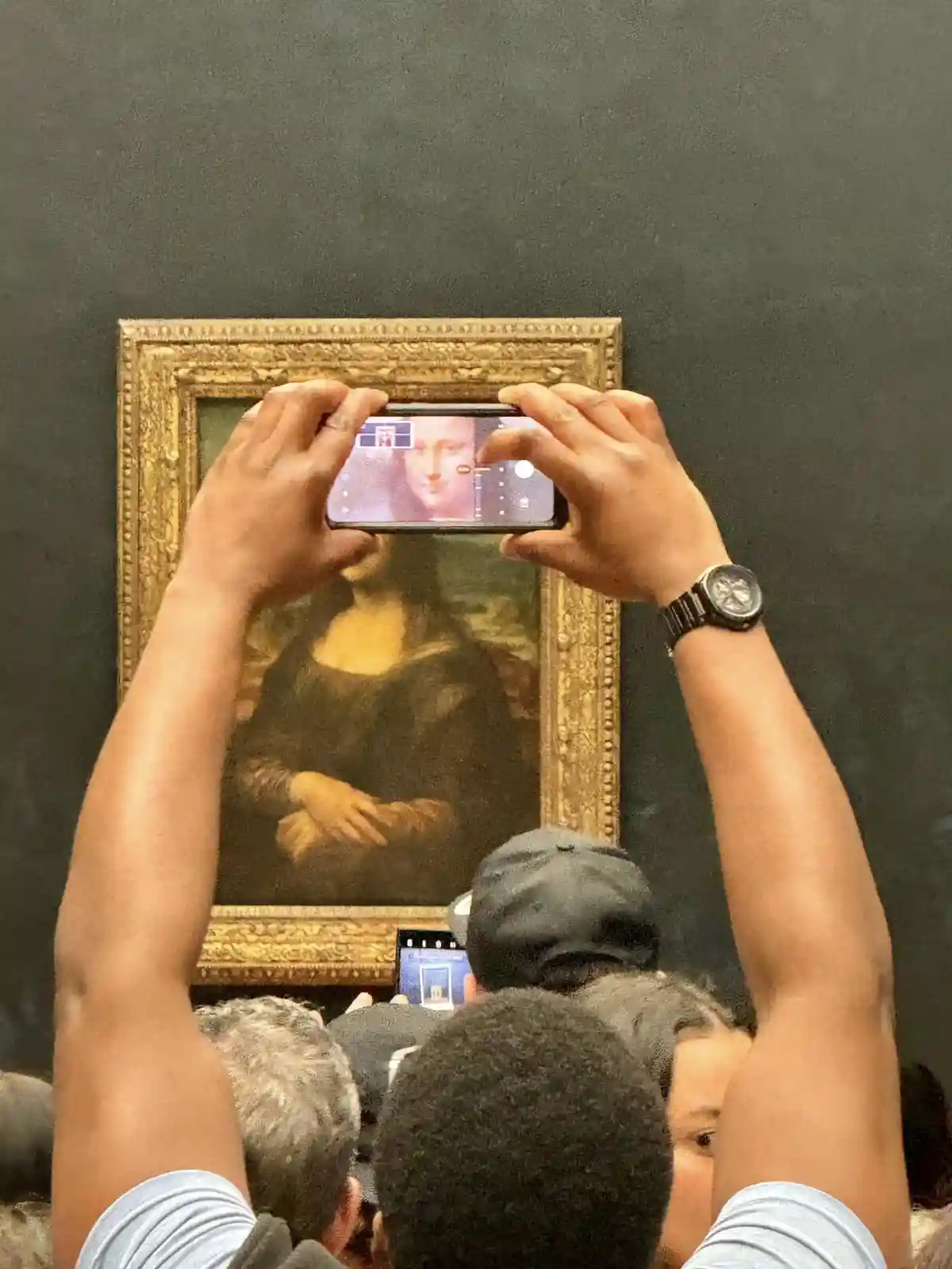 A person holds a smartphone horizontally to photograph the Mona Lisa painting, which is visible behind the phone's screen…