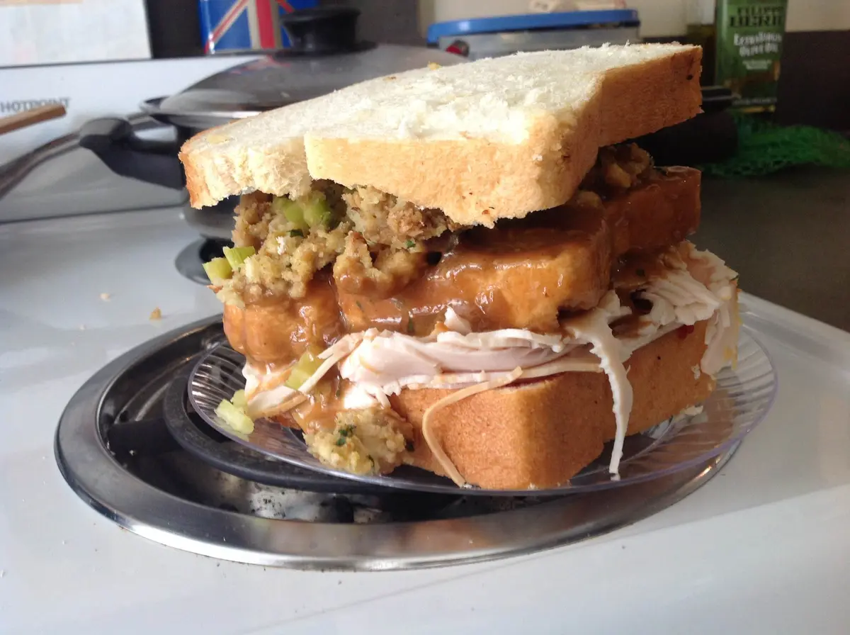 A towering, messy sandwich made with white bread, layers of shredded turkey, stuffing, gravy, and possibly bread slices…