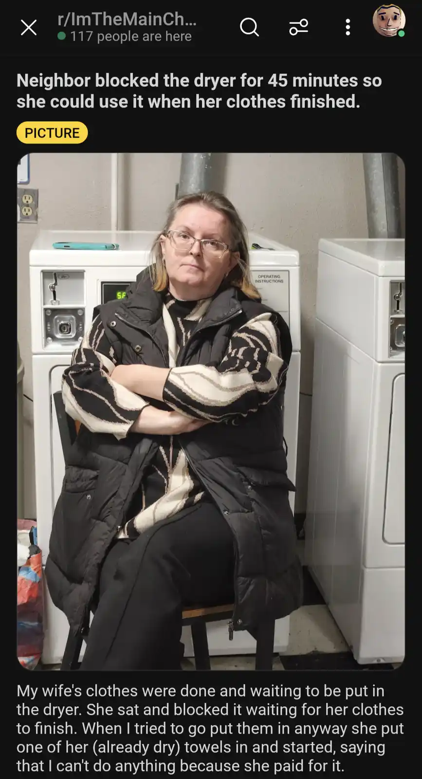 A woman with glasses and a black puffer vest sits on a chair in a laundromat, arms crossed, looking annoyed. She is…