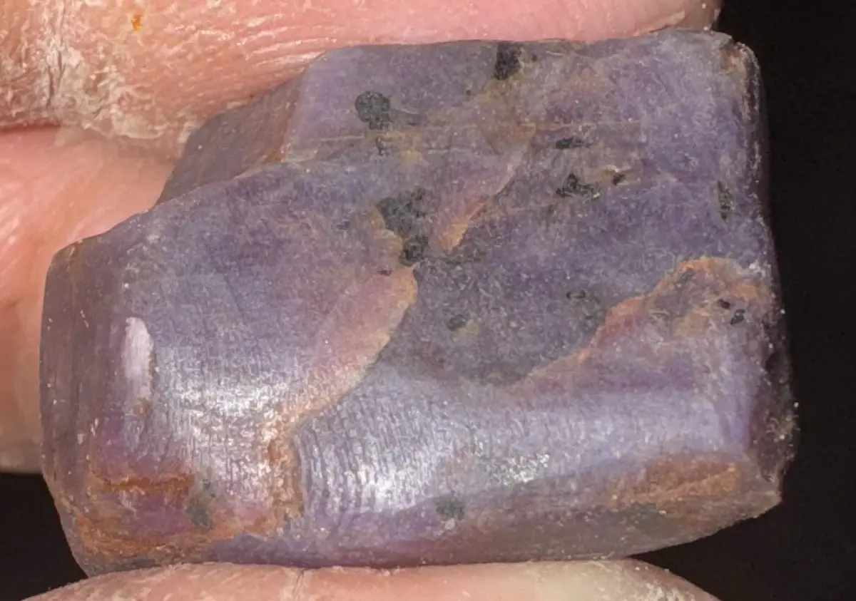 a close-up photo shows a hand holding a rough, purple amethyst crystal with visible inclusions and natural color variations…