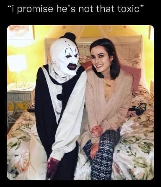 A woman with dark hair and a beige sweater sits on a floral bed next to someone in a white and black clown costume with a…