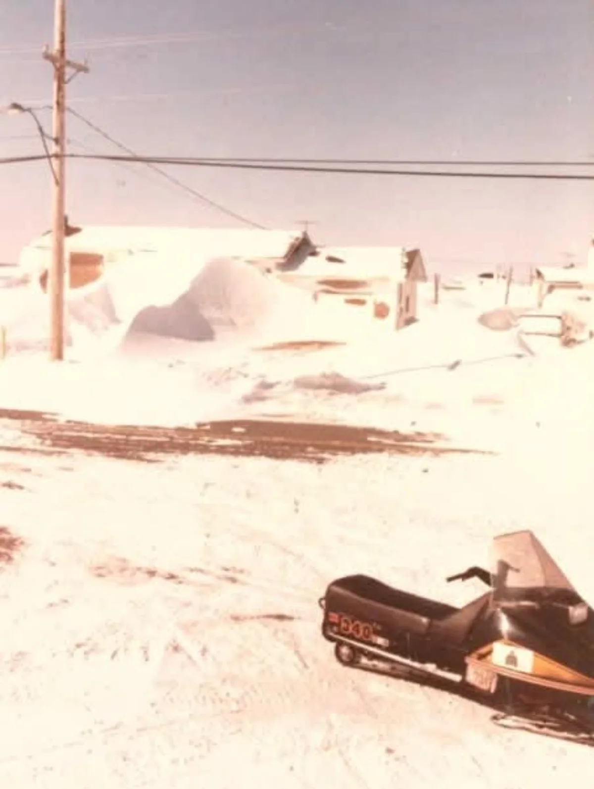 A vintage, faded photograph shows a black snowmobile parked in a snowy, rural landscape with houses and a utility pole in…