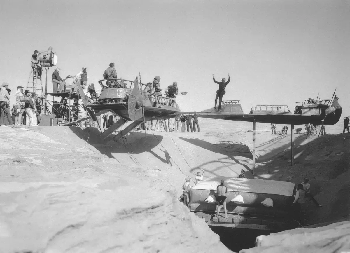 A black-and-white behind-the-scenes photo from a movie set shows a large, elaborate, and clearly impractical flying boat…