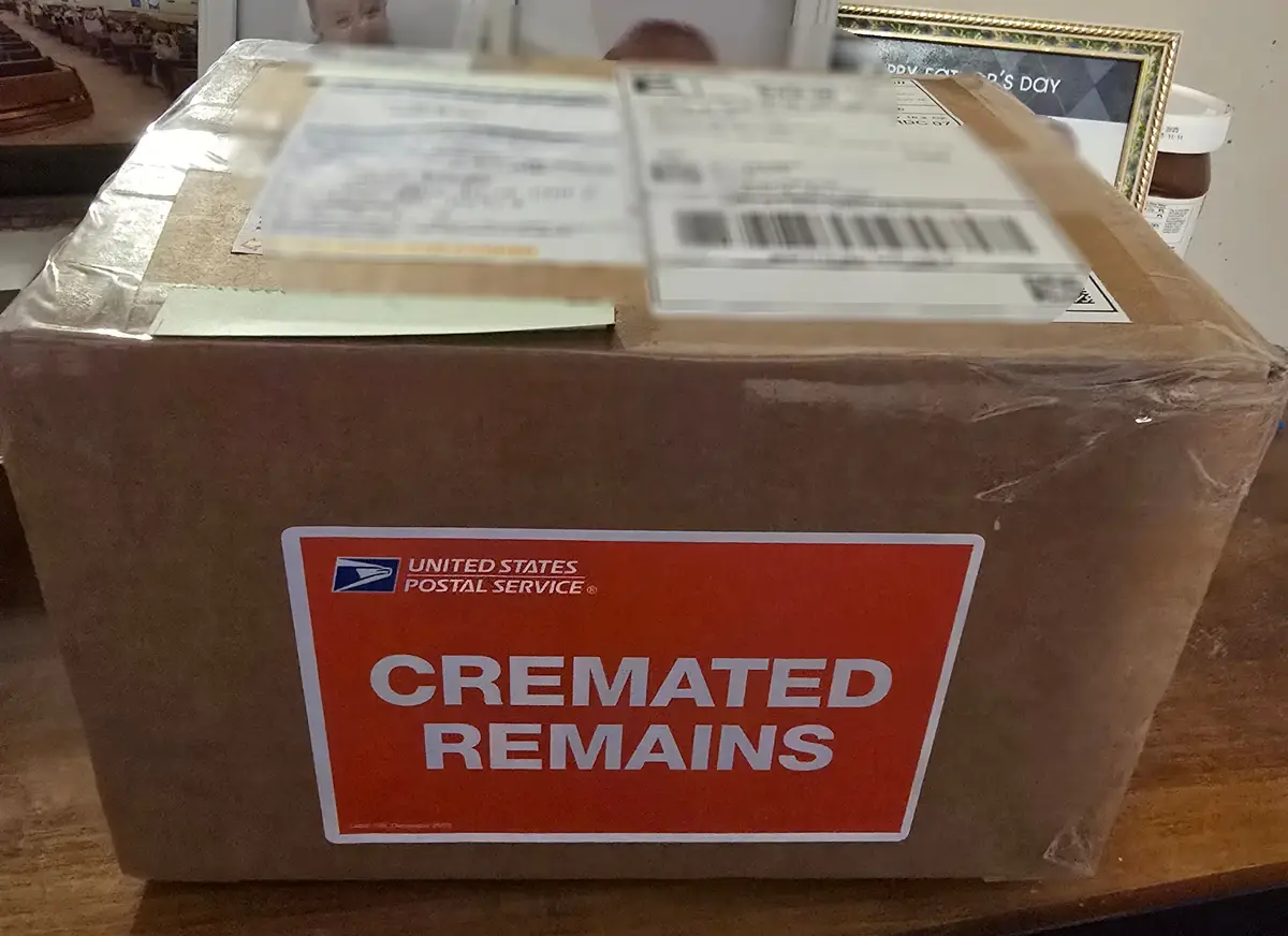 A large brown cardboard box, sealed with tape and bearing a United States Postal Service label, has a prominent red sticker…