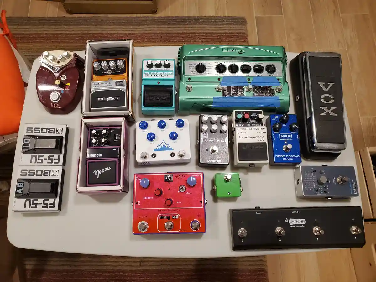 An overhead shot displays a large collection of colorful guitar effect pedals arranged on a white table, including brands…