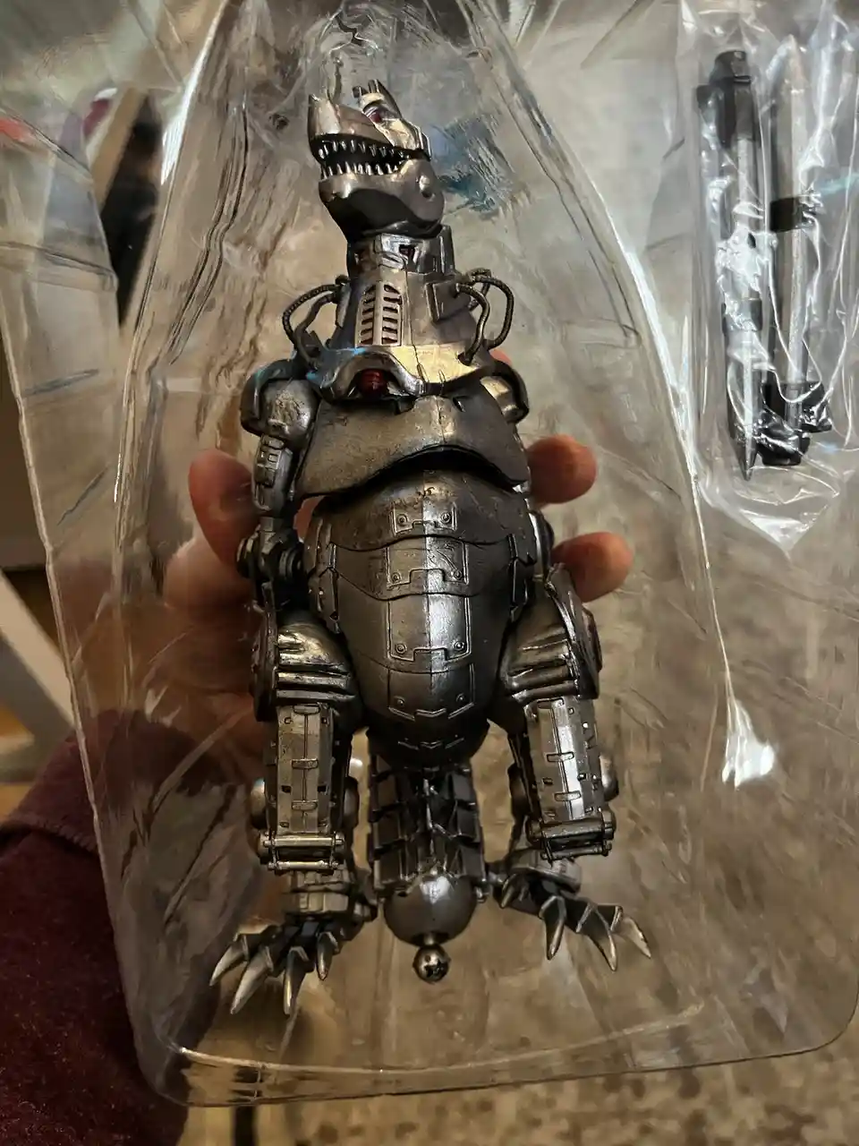A person holds a detailed, silver-colored robot dinosaur figurine encased in clear plastic packaging, with a small plastic…