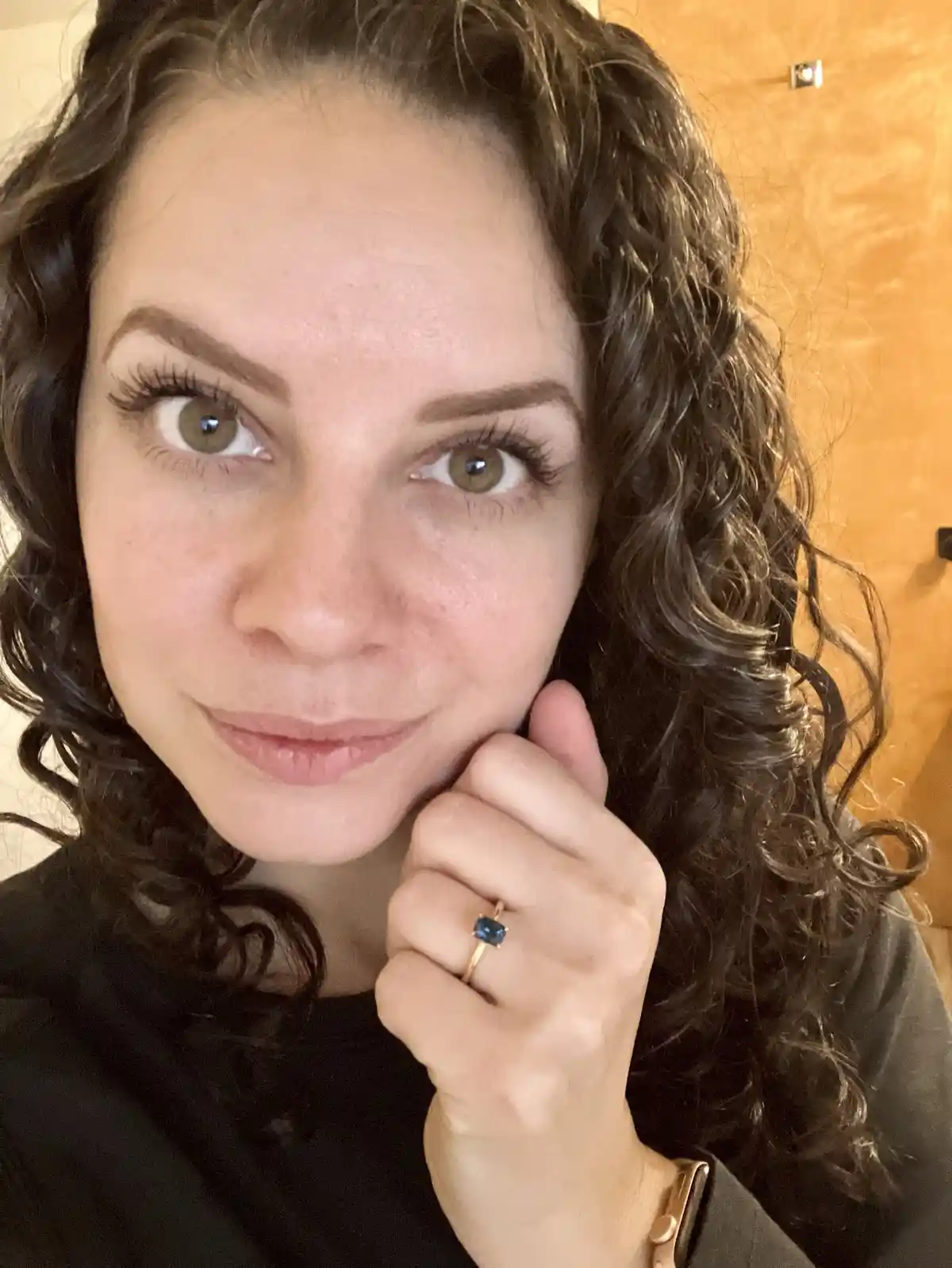 A woman with dark curly hair and green eyes looks at the camera with a slight smile while holding her hand up to her cheek…