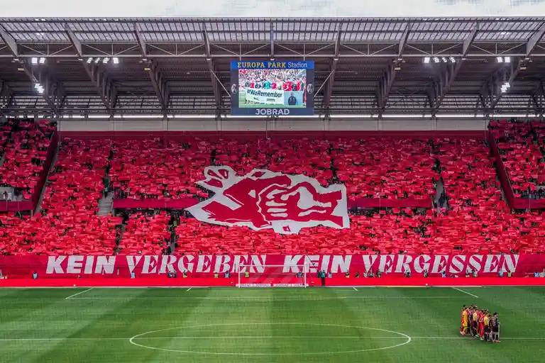 A wide shot of a football stadium filled with fans holding red banners, forming a large white graphic of a roaring lion and…