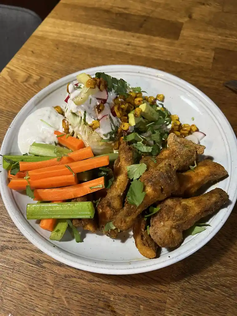 A white plate on a wooden table holds fried chicken wings, carrot and celery sticks, a creamy dip, and a colorful salad with…