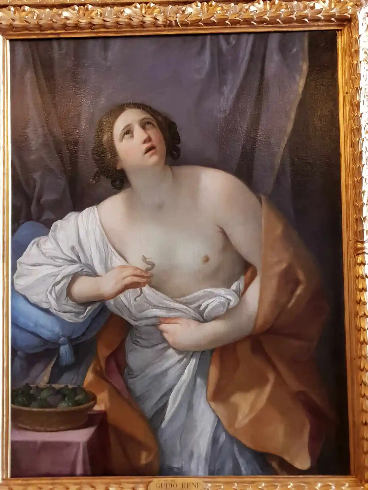 A classical painting by Guido Reni depicts a woman in flowing robes holding a small snake, with a bowl of figs nearby. The…