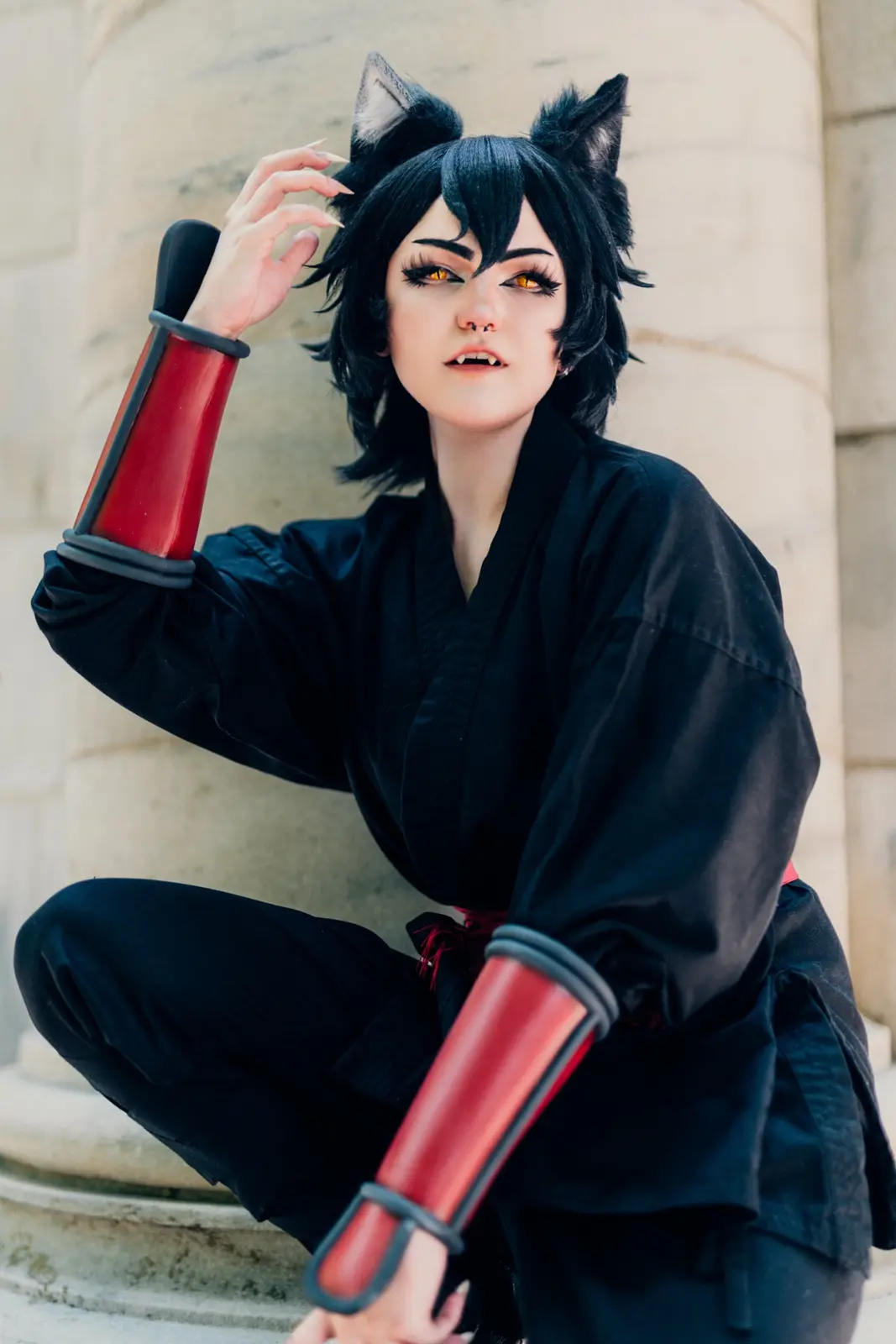 A person in a cosplay outfit with black cat ears, fangs, and yellow eyes, wearing a black kimono with red arm guards, poses…