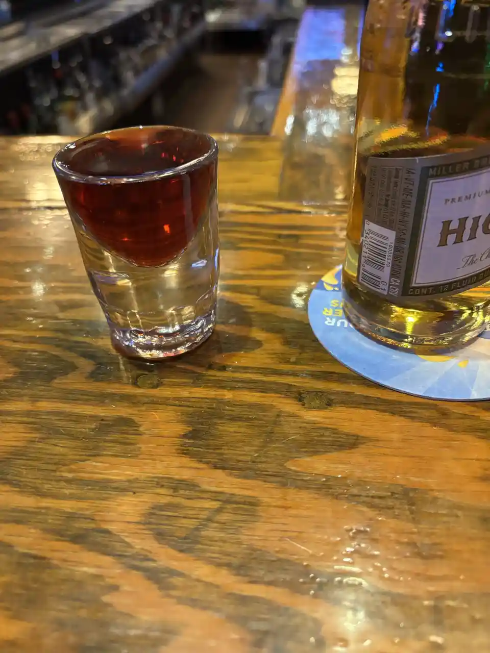 A shot glass containing a small amount of red liquid sits on a wooden bar next to a Miller High Life beer bottle. The shot…