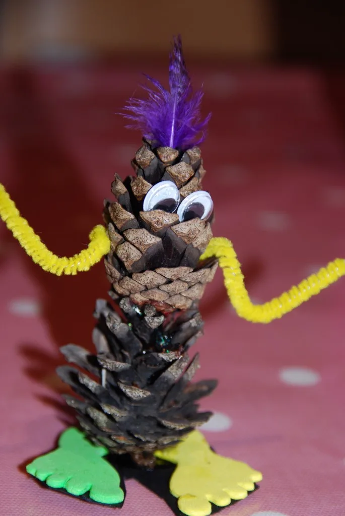 A pinecone is decorated with googly eyes, a purple feather on top, yellow pipe cleaner arms, and green and yellow foam feet…
