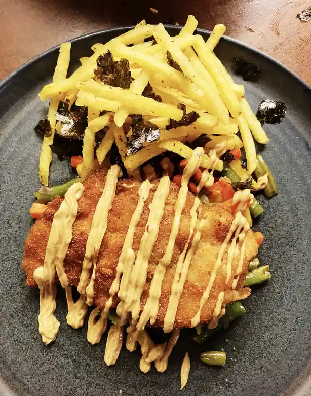 A close-up photo of a plated meal featuring a breaded and fried chicken katsu, drizzled with a creamy sauce, resting on…