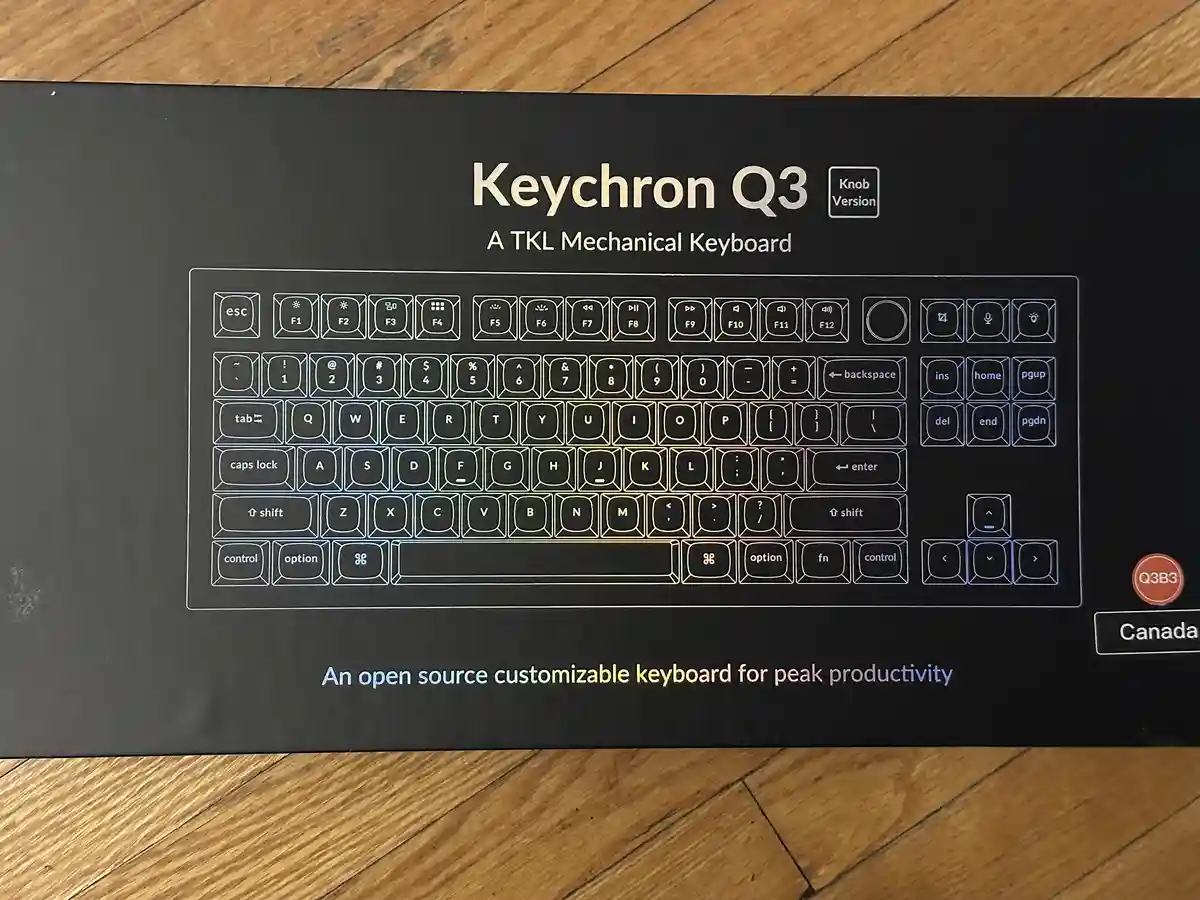 A close-up photo of a black Keychron Q3 keyboard box resting on a wooden floor. The box features a white outline of the TKL…