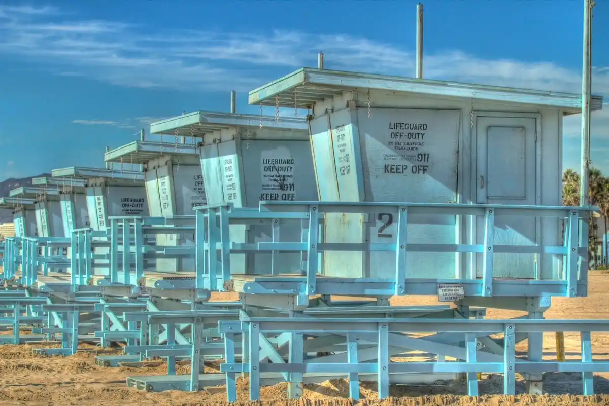 A row of empty, light-blue lifeguard towers on a sandy beach under a clear blue sky, all marked with 'LIFEGUARD OFF-DUTY'…