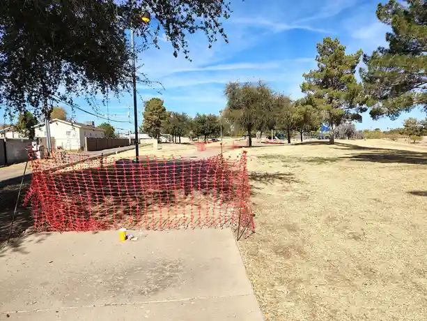 An outdoor photo shows a bright orange plastic construction fence blocking a dirt area in a dry, grassy park with trees and…