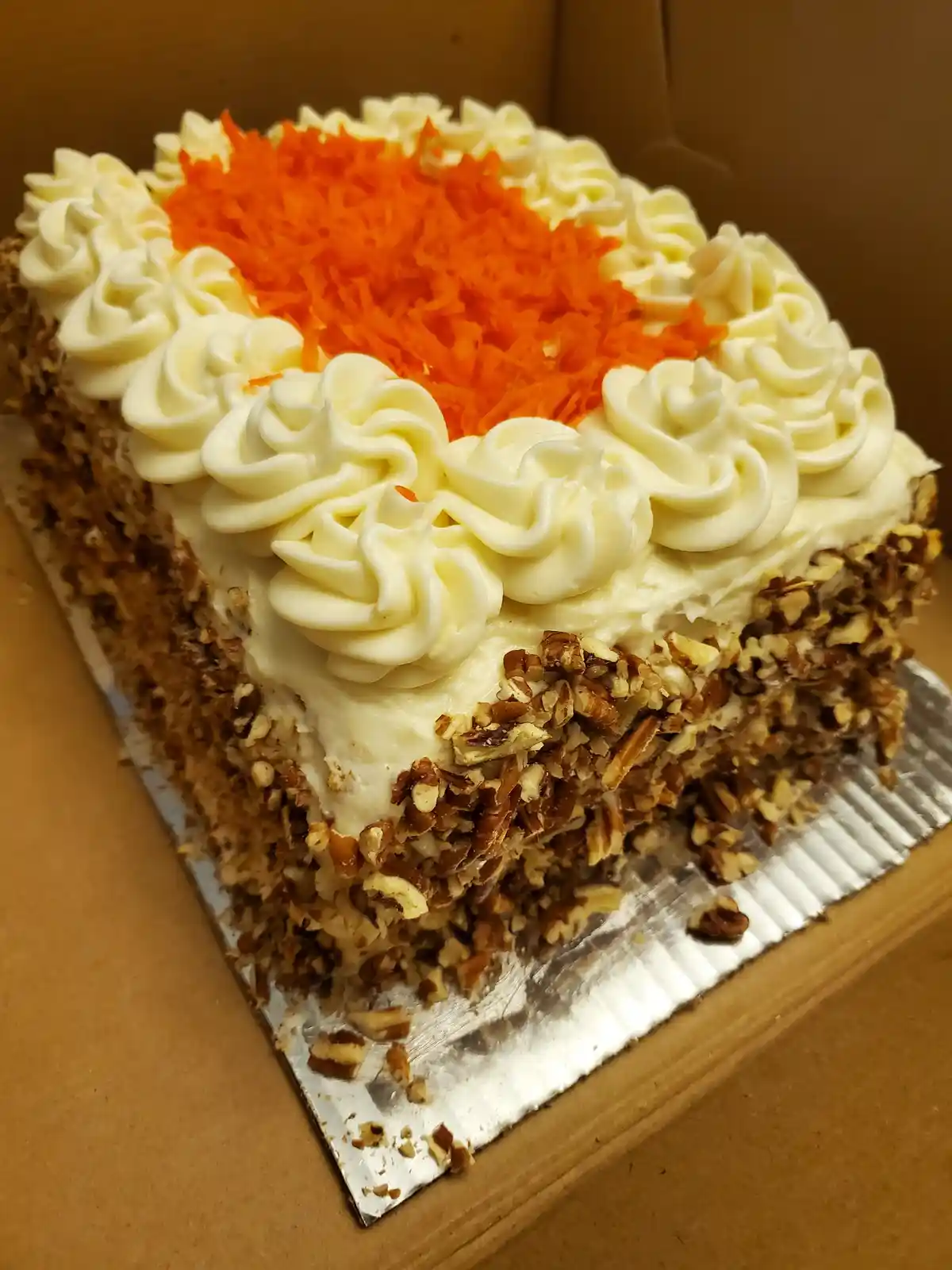 A rectangular carrot cake sits in a cardboard box on a foil-lined tray, decorated with white piped frosting swirls around…