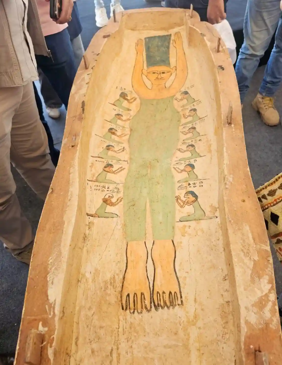 An ancient Egyptian coffin with a painted figure standing upright, arms raised, surrounded by smaller painted figures in…