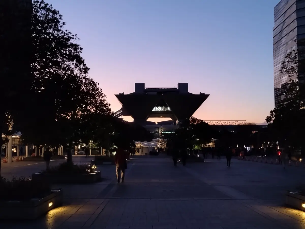 A twilight photo shows a wide plaza with silhouetted trees and people walking toward a large, distinctive building with a…