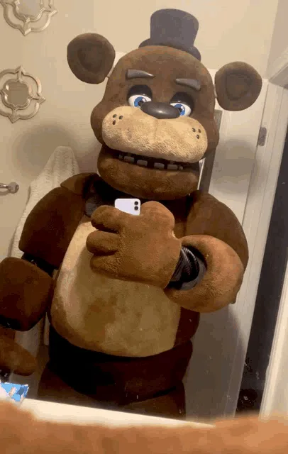 A large animatronic bear wearing a top hat and bowtie holds a smartphone to take a mirror selfie. The bear has a slightly…