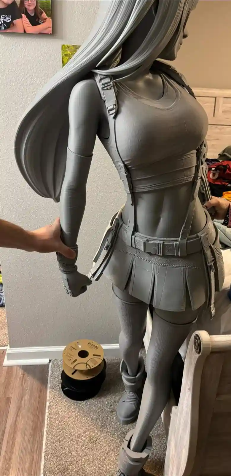 A large, grey, un-painted 3d-printed statue of an anime-style female character with long hair and tactical gear stands…