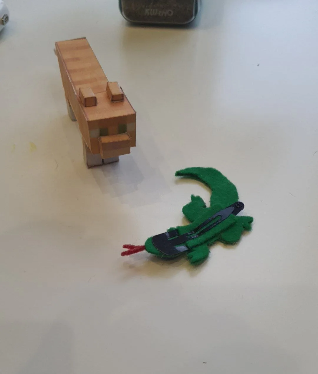 A wooden block-style Minecraft cat toy stands facing a small green felt lizard toy with a black hair clip attached, both on…