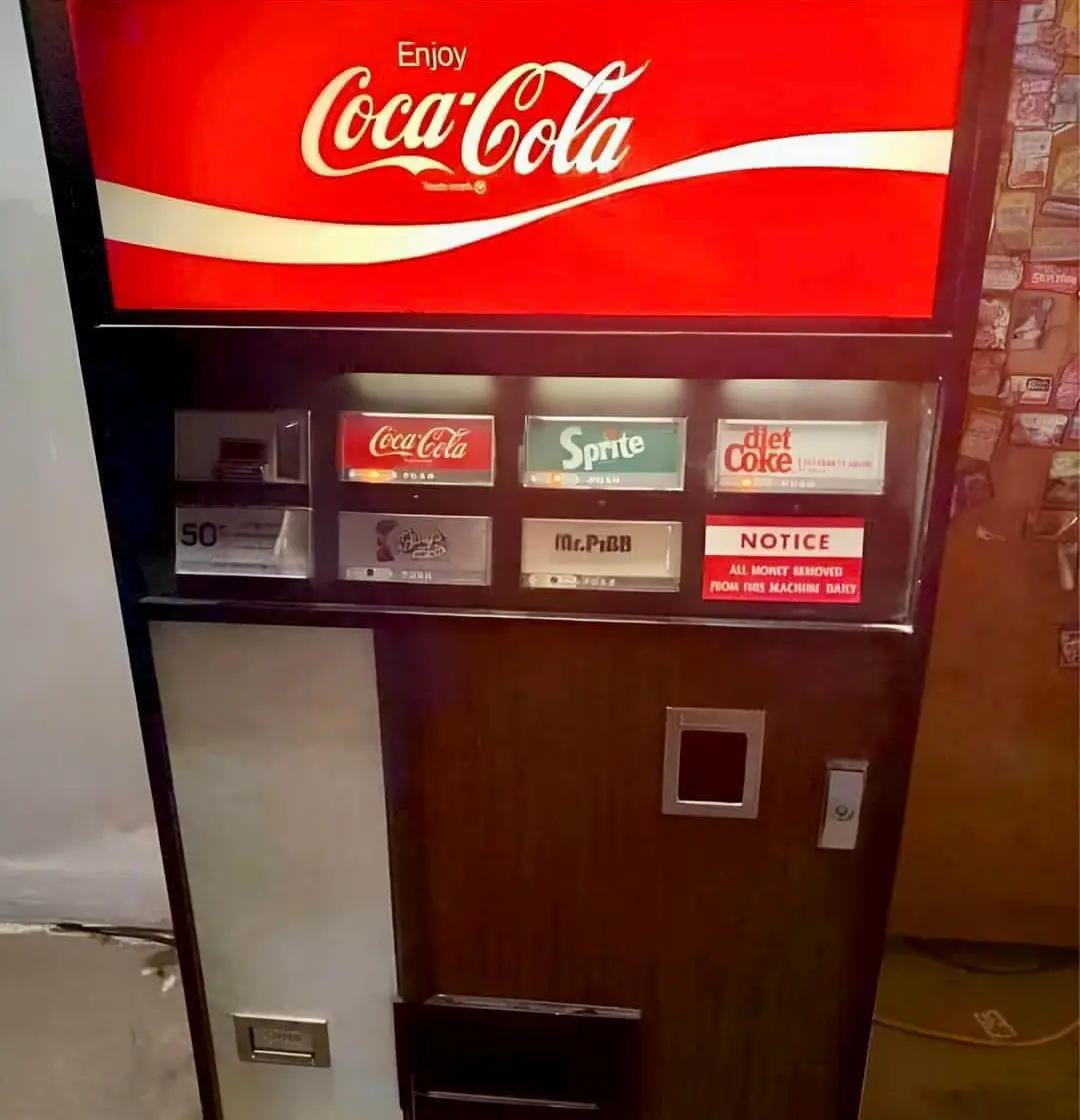 A vintage Coca-Cola vending machine displays options for Coca-Cola, Sprite, Diet Coke, and Mr. Pibb, with a red notice…