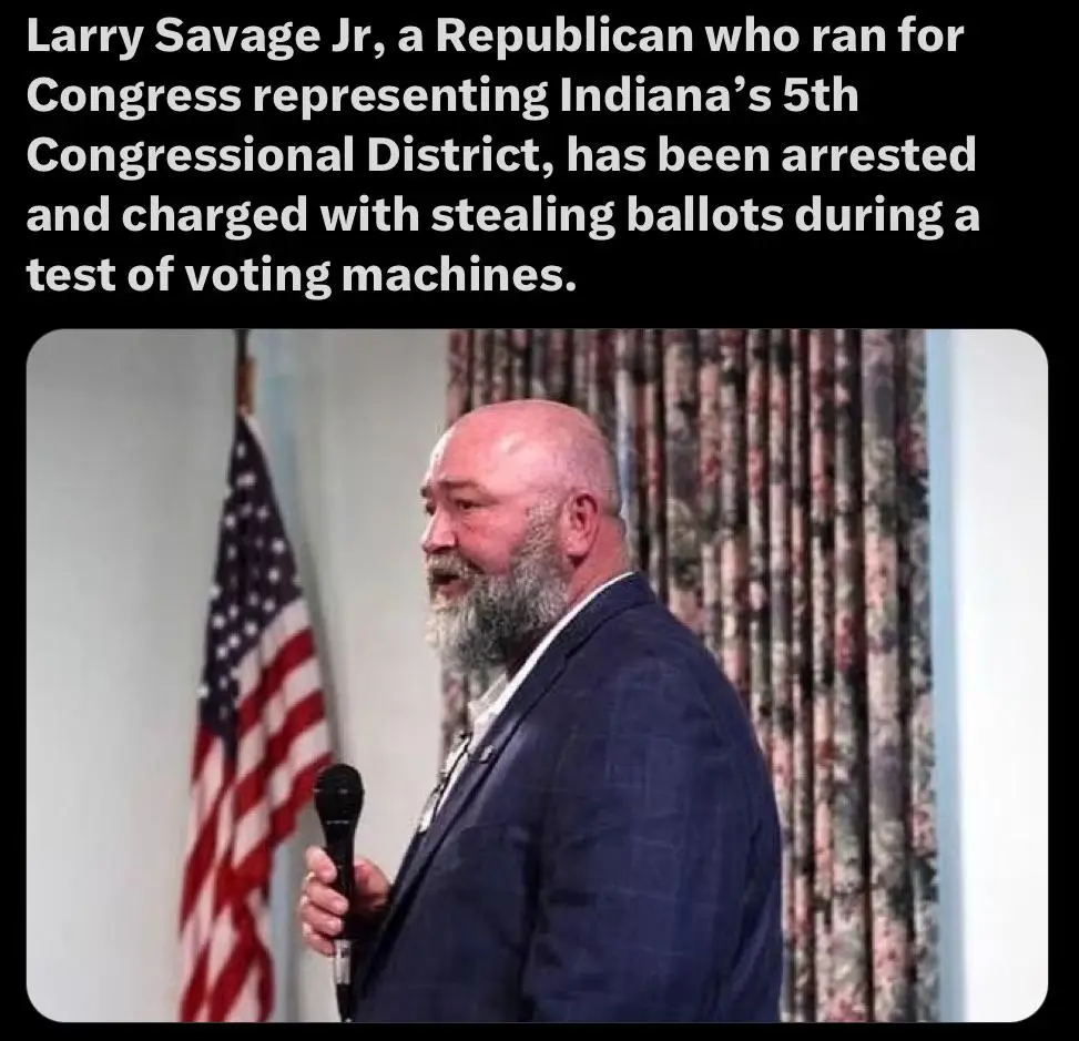 A bald man with a gray beard wearing a dark suit holds a microphone while standing in front of an American flag and…