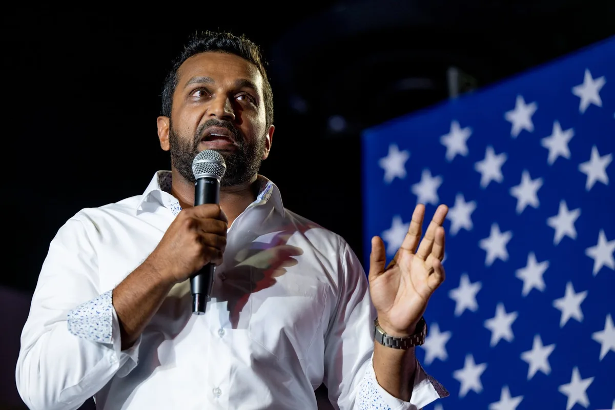 a man with a beard and dark hair, wearing a white button-up shirt with patterned cuffs, holds a microphone in one hand and…