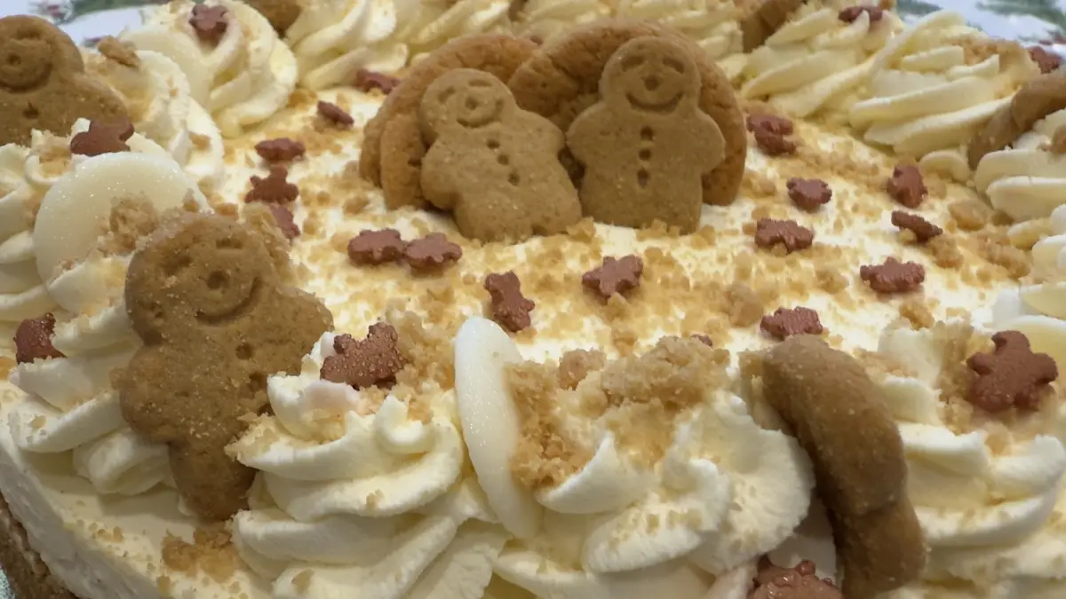 A close-up of a cake decorated with piped white cream, crumbled gingerbread, and small gingerbread man-shaped cookies…