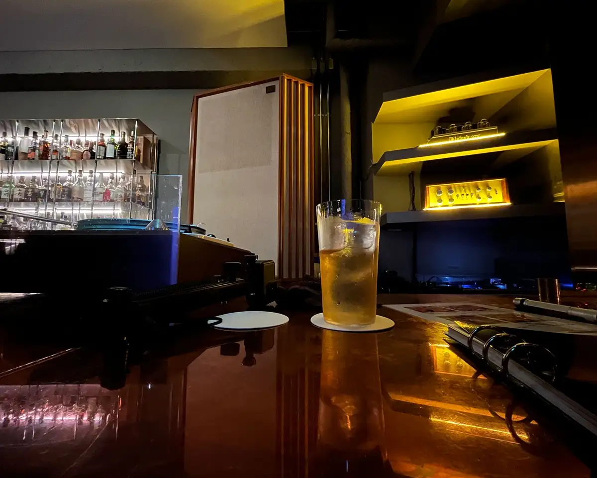 A glass of amber-colored cocktail with ice sits on a coaster on a dark, reflective bar counter. Behind it, shelves of liquor…