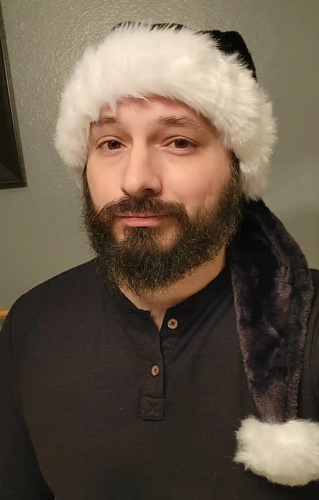 A man with a full beard and mustache wears a black and white Santa hat with a white fluffy trim and a white pom-pom, looking…