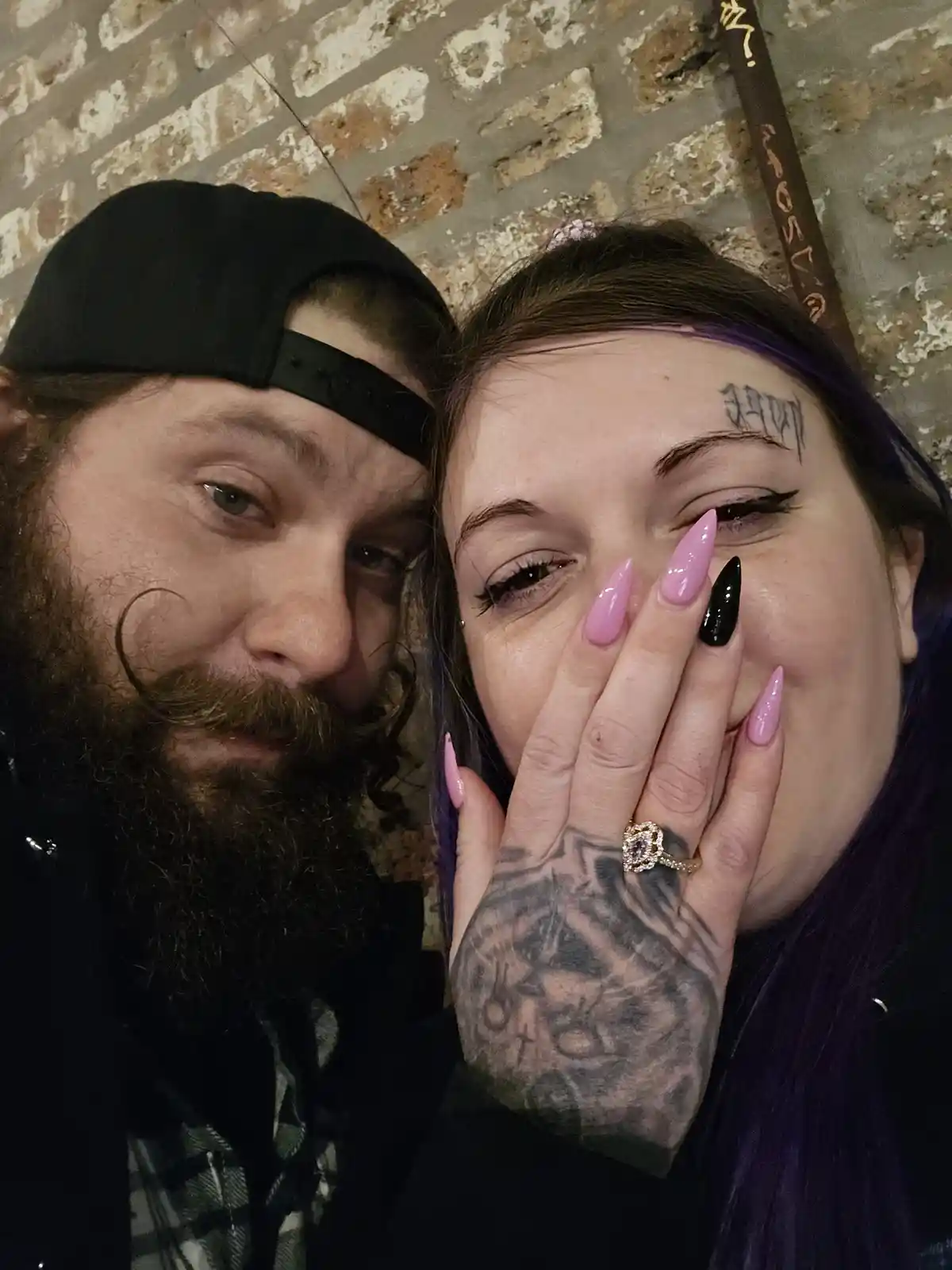 A selfie of a man with a beard and black cap next to a woman with purple hair and a tattoo on her forehead. The woman covers…